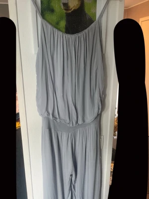 Ljusgrå slapp jumpsuit med tunna axelband - Ljusgrå jumpsuit med tunna axelband och lös passform. Plagget har ett rynkat liv och bred resår i midjan för en avslappnad look. Tillverkad i ett mjukt, lätt tyg som faller snyggt. Perfekt för dig som gillar en chill och stilren vibe.