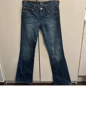 Blå bootcut jeans från 7 for all mankind - Snygga blå bootcut jeans från 7 for all mankind med klassiska fem fickor och kontrastsömmar. Jeansen har en lätt utsvängd passform nertill och är gjorda i denim med en autentisk tvätt. Perfekta för en avslappnad och trendig look. Dragkedjan är sönder därav priset!