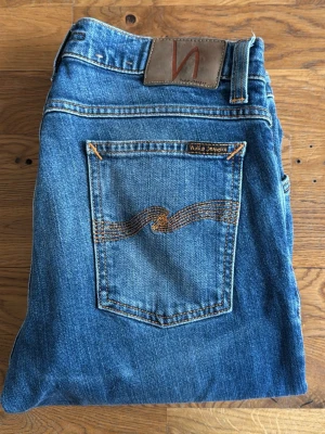 Blå jeans från Nudie Jeans - Säljer ett par klassiska blå jeans från Nudie Jeans med snyggt slitna detaljer och orange söm på bakfickan. Jeansen har rak passform. Använt dem max 2 gånger inga tecken på slitage.