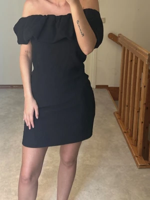 Svart offshoulder kort klänning - Trendig svart kort klänning med offshoulder och volang upptill. Klänningen har en figurnära passform och är perfekt för dig som gillar en stilren look. Materialet känns mjukt och bekvämt. 