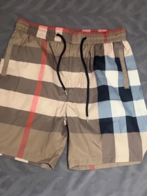 Rutig shorts med snörning - Snygga shorts med klassiskt rutigt mönster i beige, brunt, blått och rött. Shortsen har elastisk midja med svart snörning och sidofickor. Perfekta för sommaren och ger en trendig look.