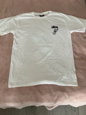 Vit Stüssy t-shirt med tärningsprint - Vit t-shirt från Stüssy med svart logga och två tärningar tryckta på bröstet. Klassisk passform och rund halsringning. Perfekt för dig som gillar streetwear och vill ha en clean look med en twist.