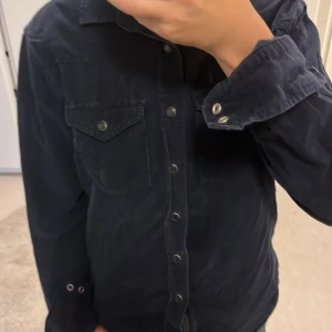 Mörkblå manchester overshirt - Säljer en mörkblå overshirt i mjuk manchester med tryckknappar framtill och två bröstfickor med lock. Skjortan har klassisk krage och långa ärmar med knappdetaljer vid manschetterna. Perfekt för lager-på-lager och chill stil. Passar ungdomar under 160.