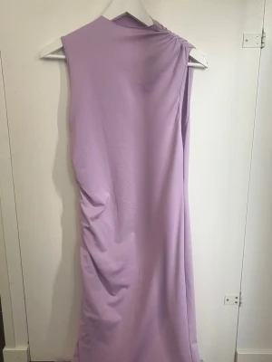 Ljuslila långklänning med drapering - Säljer en elegant långklänning i ljuslila med draperad detalj vid axeln och sidan. Klänningen är ärmlös och har en mjuk, följsam passform som ger en snygg siluett. Perfekt för dig som vill sticka ut med en stilren look.