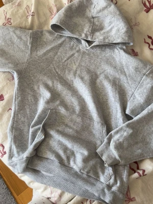 Grå basic hoodie med ficka - En enkel och stilren grå hoodie med huva och stor magficka. Hoodien har ribbade muddar vid ärmslut och nederkant. Perfekt för en avslappnad look och passar till det mesta. Materialet känns mjukt och bekvämt mot huden.