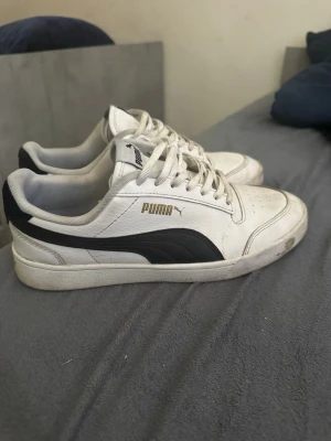 Vita Puma sneakers med svart detalj - Snygga vita sneakers från Puma med klassisk svart sidodetalj och logga i guld. Skorna har lågt skaft, vita snören och texten 'Since 1948' på plösen. Perfekta för en stilren och sportig look. Ovandelen är i syntetmaterial och textil.