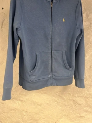 Blå zip hoodie från Ralph Lauren - Blå hoodie med dragkedja från Ralph Lauren. Klassisk design med två fickor framtill och gul broderad logga på bröstet. Mjuk bomullskänsla och ribbade muddar vid ärmslut och nederkant. Perfekt för en avslappnad och stilren look.