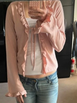 Ljusrosa volangkofta med knytband - Supersöt ljusrosa kofta med volangkant längs öppningen och ärmsluten. Knyts framtill med smala band och har en croppad passform. Perfekt att slänga över ett linne för en gullig look.
