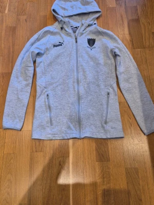 Ljusgrå Malmö FF hoodie från Puma - Ljusgrå hoodie från Puma med Malmö FF-logga på bröstet. Dragkedja framtill, två fickor med dragkedja och huva. Tillverkad i mjukt material som känns skönt mot huden. Perfekt för dig som vill representera ditt lag med stil.