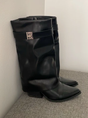 Svarta boots med metallspänne - Säljer ett par svarta boots i mjukt skinn med coolt metallspänne på sidan. Stövlarna har en slouchy passform, spetsig tå och låg klack. Skinnet har ingen skada det är endast lite smuts på klacken som såklart tas bort vid försäljning. Använd 2 gånger, jätte bekväma, storlek 37 1/5