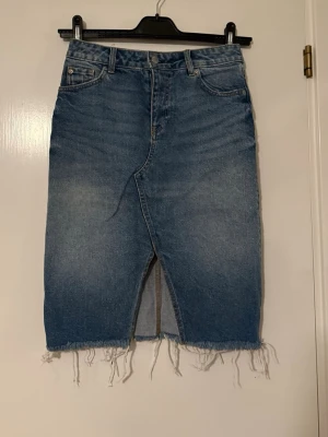 Blå jeanskjol med rå kant från H&M - Trendig blå jeanskjol från H&M i storlek 34. Kjolen har en rak passform, hög midja, klassiska fem fickor och en snygg slits framtill. Rå, fransig kant nedtill ger en cool och avslappnad vibe. Perfekt att matcha med sneakers eller boots.