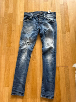 Slim fit jeans från Jack & Jones - Snygga blå slim fit jeans från Jack & Jones med slitningar och hål på ena benet. Jeansen har klassisk femficksdesign, knappgylf och ljusa tvättade partier för en trendig look. Perfekta för dig som gillar en avslappnad och modern stil.