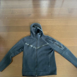Svart Nike hoodie med dragkedja - Snygg svart hoodie från Nike med hel dragkedja och huva. Har en diskret logga på bröstet och en cool diagonal detalj över bröstet. Ficka med dragkedja på ena ärmen. Perfekt för en sportig och avslappnad stil. Sitter så på mig som är 180 så kanske passar en L också