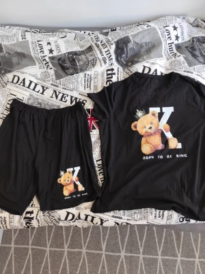 Svart pyjamas med nalletryck - Mysig svart pyjamas med t-shirt och shorts. Båda delarna har ett gulligt tryck av en nalle med krona och texten 'Born to be king' samt ett stort K. Perfekt för chillkvällar hemma. Stor i storleken 