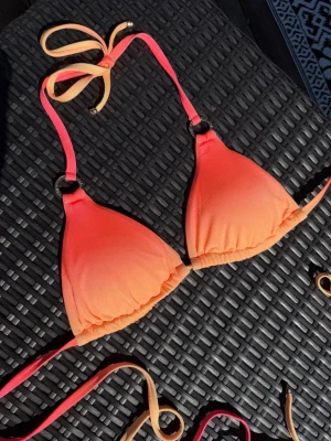 Ombre triangelbikini i orange och rosa - Snygg triangelbikini med ombre-effekt i orange och rosa nyanser. Bikinitoppen har tunna knytband i nacken och ryggen samt små ringdetaljer vid axelbanden. Banden runt ryggen är extra långa för att kunna knyta flera varv runt magen!🩷🧡