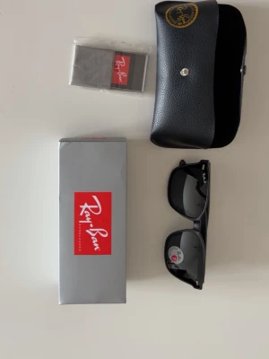 Svarta Ray-Ban Wayfarer solglasögon - Klassiska svarta Wayfarer solglasögon från Ray-Ban med ikonisk fyrkantig form och mörka glas. Kommer med originalfodral, putsduk och box. Perfekta för att lyfta din stil med en tidlös accessoar.