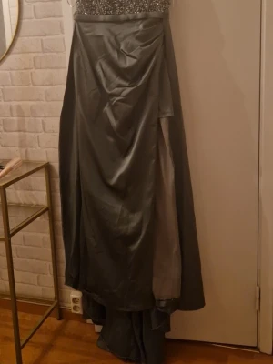 Lång grå balklänning med paljetter - Elegant långklänning i oliv grön med glittrig paljettliv och svepande kjol i satinliknande material. Klänningen har axelbandslös design och vacker drapering framtill, perfekt för bal eller festliga tillfällen.ej kemtvättat 