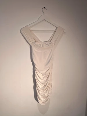 Vit omlott kortklänning med drapering - Supersnygg vit kortklänning med breda axelband och omlott-detalj. Klänningen har draperad effekt längs sidorna som ger en cool och trendig look. Perfekt för dig som vill sticka ut med en elegant men modern stil.