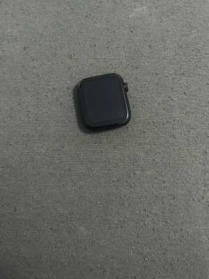 Svart Apple Watch smartklocka - En Snygg svart 7 series Apple Watch med fyrkantig form och digital krona på sidan. Klockan har en slät glasfront och baksida med sensorer för pulsmätning. Perfekt för dig som vill ha koll på hälsa och notiser direkt på handleden.