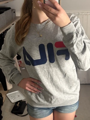 Grå sweatshirt från FILA - Snygg grå sweatshirt från FILA med stort blått och rött logotryck på bröstet. Tröjan har rund halsringning, långa ärmar och ribbade muddar vid ärmslut och nederkant. Hör av er om frågor 