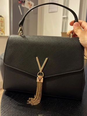 Valentino handväska - Stilren svart handväska från Valentino med guldfärgade detaljer och kedjor. Väskan har ett elegant V-spänne framtill och ett avtagbart handtag. Tillverkad i strukturerat konstläder med klassisk, fyrkantig form. Nästan nyskick.