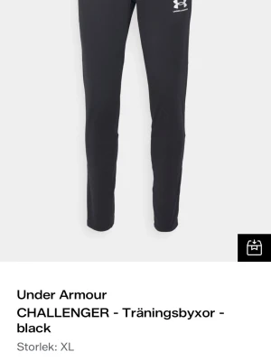 Under Armour svarta träningsbyxor XL - Svarta träningsbyxor från Under Armour i modellen CHALLENGER. Byxorna har smal passform, elastisk midja och vitt logotryck på låret. Perfekta för träning och sportaktiviteter. Tillverkade i ett lätt och funktionellt material.