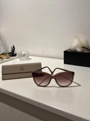 Bruna cat eye-solglasögon - Snygga solglasögon i cat eye-modell med brunt transparent plastbåge och tonade bruna glas. Kommer med ett stilrent beige fodral med logga. 