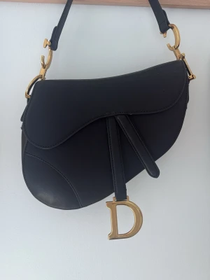 Svart Dior Saddle Bag i läder - Ikonisk svart Dior Saddle Bag i slätt läder med guldfärgade metalldetaljer och D-formad hängande charm. Väskan har en unik sadelform och justerbar axelrem med C-formade spännen. Perfekt statement piece för dig som gillar lyxiga accessoarer.