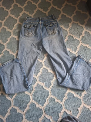 Blå bootcut jeans med broderade fickor - Snygga ljusblå bootcut jeans med broderade detaljer och knappar på bakfickorna. Jeansen har klassisk femficksdesign och är tillverkade i denim. Perfekta för dig som gillar en avslappnad men ändå trendig stil. Low waist/mid waist