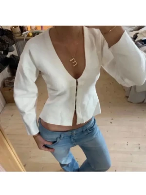 Vit v-ringad blus med knappar - Snygg vit blus med v-ringning och knappar framtill. Modellen är croppad och har långa ärmar. Perfekt att styla med jeans eller kjol för en trendig look.