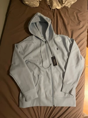 Ljusblå hoodie från Polo Ralph Lauren - Snygg ljusblå hoodie från Polo Ralph Lauren med dragkedja framtill, huva med dragsko och klassisk logga broderad på bröstet. Tillverkad i mjuk bomullsblandning och har två fickor framtill. Perfekt för en chill och stilren look.