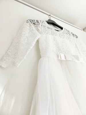 Vit elegant klänning för flicka  - Elegant vit långklänning med spets upptill, trekvartsärm och en söt rosett i midjan. Kjolen är gjord i flera lager tunn tyll som ger en drömmig känsla. Perfekt för speciella tillfällen där du vill känna dig extra fin.