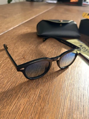 Moscot Lemtosh 46 - Snygga svarta solglasögon från Lemtosh med rundad form och blåtonade glas. Bågarna är i blank svart plast och kommer med ett stilrent svart fodral. Perfekta för dig som vill ha en klassisk men trendig look.