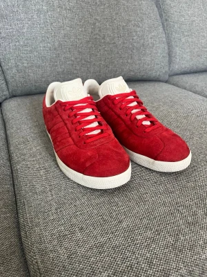 Röda Adidas Gazelle sneakers i mocka - Snygga röda Adidas Gazelle sneakers i mjuk mocka med vita detaljer och klassisk vit sula. Skorna har lågt skaft, röda skosnören och ikonisk Adidas-logga på plösen. Perfekta för dig som vill sticka ut med färg och stil.