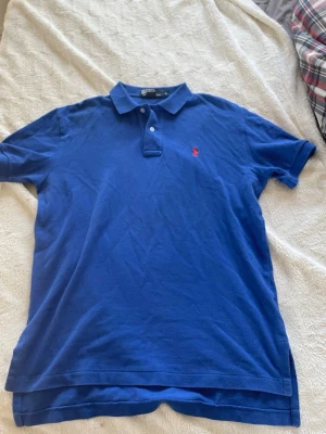 Blå pikétröja från Ralph Lauren - Blå pikétröja från Ralph Lauren med klassisk krage, två knappar vid halsen och röd broderad logga på bröstet. Tillverkad i mjuk bomull och har korta ärmar. Storlek s. Perfekt inför en varm sommardag ☀️ ✨👕