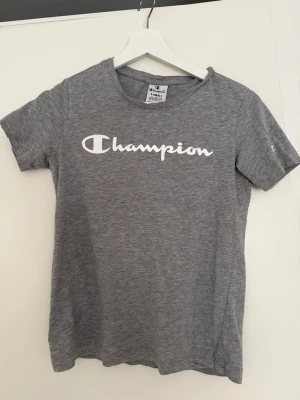 Grå Champion t-shirt med logga - Snygg grå t-shirt från Champion med klassisk vit logga tryckt över bröstet. T-shirten har rund halsringning och korta ärmar. Tillverkad i mjuk bomull som känns skön mot huden. Perfekt till jeans eller shorts för en avslappnad stil.