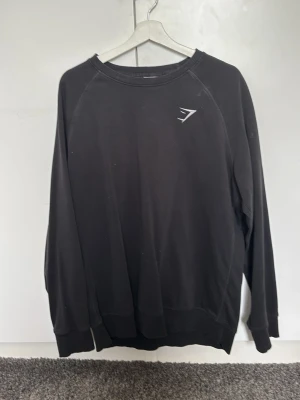 Svart sweatshirt från Gymshark - Svart sweatshirt från Gymshark med rund halsringning och diskret logga på bröstet. Tröjan har långa ärmar och ribbade muddar vid ärmslut och nederkant. Perfekt för en avslappnad och sportig stil. Det är XL men mer åt L