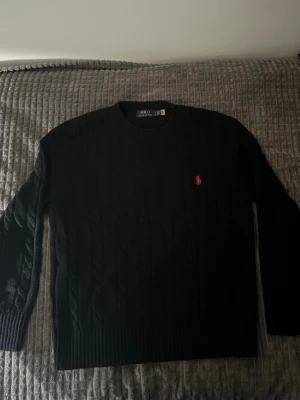 Svart kabelstickad tröja Polo Ralph Lauren - Svart kabelstickad tröja från Polo Ralph Lauren med rund halsringning och röd broderad logga på bröstet. Tröjan har långa ärmar och ribbade muddar vid ärmslut och nederkant. Perfekt för en stilren och klassisk look.