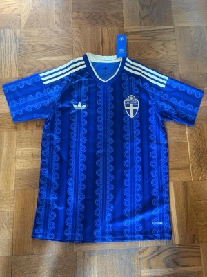 Blå Sverige fotbollströja Adidas - Säljer en blå fotbollströja från Adidas med Sveriges landslagsemblem på bröstet. Tröjan har vita detaljer, klassiska Adidas-ränder på axlarna och ett unikt mönster över hela tyget. Perfekt för dig som vill stötta Sverige med stil.