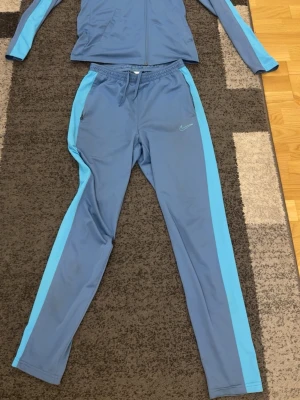 Blått träningsset från Nike (Nike dri fit ) - Snyggt blått träningsset från Nike med jacka och byxor. Setet har ljusblå ränder längs ärmar och ben samt Nike-logga på byxan. Tillverkat i ett mjukt och stretchigt material som passar perfekt för träning eller sport passar även xs