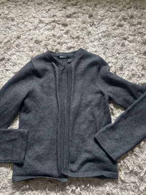 Mörkgrå kofta från Gina Tricot - Mörkgrå kofta från Gina Tricot med rund halsringning och knappstängning framtill. Koftan har långa ärmar och ribbade kanter vid hals, ärmslut och nederkant. Perfekt att slänga över en t-shirt eller linne.