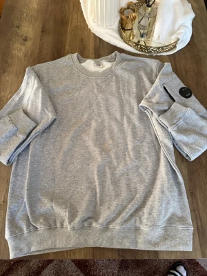 C.P. Company sweatshirt  - Säljer en stilren grå sweatshirt från C.P. Company med rund hals och ribbade muddar. Tröjan har en ikonisk svart linsdetalj på ena ärmen och är tillverkad i mjuk bomullsblandning. Perfekt för en avslappnad och trendig look.