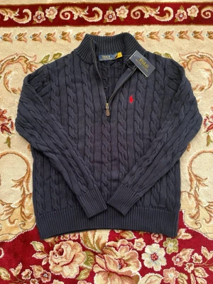 Mörkblå kabelstickad tröja Polo Ralph Lauren - Kabelstickad mörkblå tröja från Polo Ralph Lauren med halv dragkedja och ribbade muddar. Tröjan har en klassisk röd logga broderad på bröstet och är tillverkad i mjukt material. Perfekt för lager-på-lager och snygg till jeans.