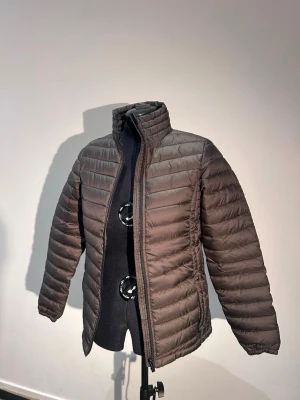 Brun pufferjacka med ståkrage - Snygg brun pufferjacka med horisontella sömmar och hög ståkrage. Jackan har dragkedja framtill och är lätt vadderad för extra värme. Perfekt för kalla dagar och enkel att matcha med olika outfits.