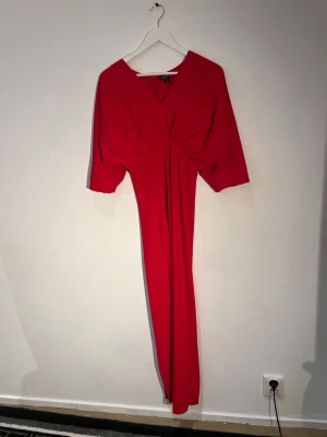 Röd långklänning med v-ringning - Snygg röd långklänning med v-ringad hals och vida trekvartsärmar. Klänningen har en draperad detalj framtill som ger en cool och trendig look. Perfekt för dig som vill sticka ut med en färgstark och elegant stil.