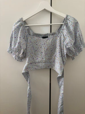 Blommig croppad blus med puffärm - Supersöt ljusblå croppad blus med små blommor i olika färger. Blusen har puffiga korta ärmar, fyrkantig ringning och knytband i midjan. Perfekt för en somrig look och passar till både jeans och kjol.