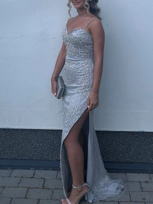 Silverglittrig långklänning med slits - Elegant långklänning i silver med tunna axelband och hög slits. Använd endast en gång. Klänningen har tappat några diamanter.