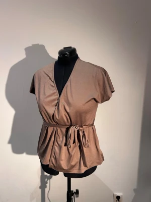 Beige omlottblus med knytskärp - Snygg beige omlottblus med korta ärmar och knytskärp i midjan. Blusen har en djup v-ringning och är tillverkad i ett mjukt, lätt material som ger ett avslappnat men stilrent intryck.