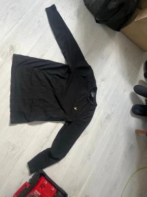 Svart långärmad topp från Lyle & Scott - Svart långärmad topp från Lyle & Scott med klassisk rund halsringning och den ikoniska gula logon broderad på bröstet. Tillverkad i mjuk bomull som är skön att bära. Enkel och stilren design som passar till det mesta.