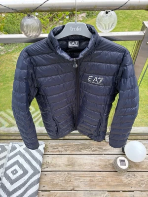 Mörkblå pufferjacka från EA7 Emporio Armani - Snygg svart pufferjacka från EA7 Emporio Armani med diskret logga på bröstet. Jackan har ståkrage, dragkedja framtill och quiltad design. Perfekt för kalla dagar och ger en sportig vibe. Materialet är glansigt och lätt, vilket gör den enkel att bära. Jacka är väldigt luftig och perfekt nu när de börjar bli varmare och lite kalla mornar, skriv vid minsta fundering🙌🏻🙌🏻